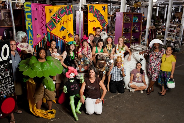 A group in odd and colorful outfits pose in front of the cluttered shelves of the Scrap Exchange